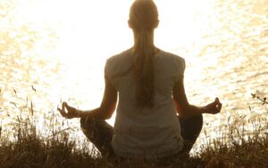 Mujer meditando en la orilla del agua al atardecer con luz dorada, en posición de loto con gesto de calma y equilibrio