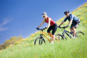 Pareja con casco montando en bicicleta de montaña por una ladera verde bajo un cielo azul