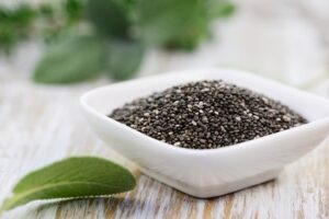 Cuenco blanco lleno de semillas de chía sobre mesa de madera con hojas de salvia
