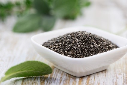 Cuenco blanco lleno de semillas de chía sobre mesa de madera con hojas de salvia