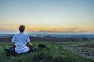 Joven meditando sentado en la hierba de una colina frente a un paisaje al atardecer
