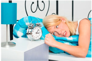Mujer rubia en la cama con gesto de insomnio junto a un despertador y una lámpara azul