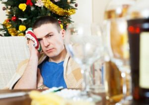 Hombre con resaca sujetándose la cabeza junto a copas y un árbol de Navidad al fondo