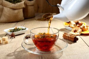 Tetera blanca sirviendo té en taza de cristal con especias, frutas secas y hierbas sobre mesa de madera