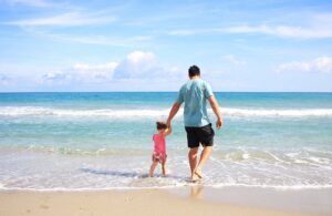 Padre e hija de la mano caminando hacia el mar en la orilla de una playa soleada