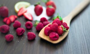 Frambuesas en cuchara de madera junto a fresas, lima y azúcar sobre mesa oscura