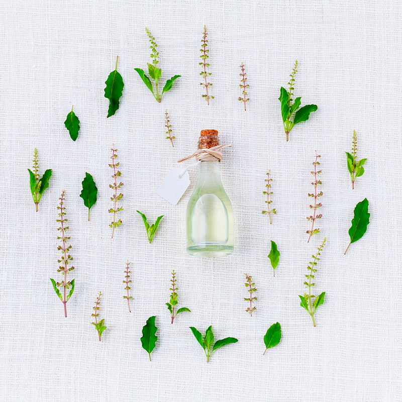 Botella de aceite esencial con etiqueta rodeada de hojas y flores de albahaca sobre lino blanco