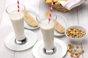 Dos vasos de horchata con pajitas rojas junto a fartons y un cuenco de chufas sobre mesa blanca