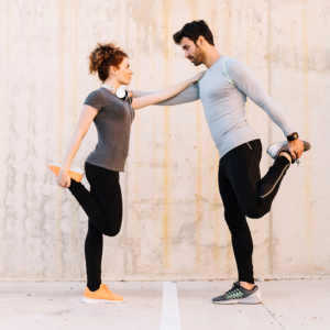 Pareja deportista estirando el cuádriceps apoyándose mutuamente frente a un muro de hormigón