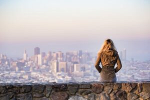 Mujer rubia sentada en un muro de piedra contemplando el skyline de una ciudad al atardecer