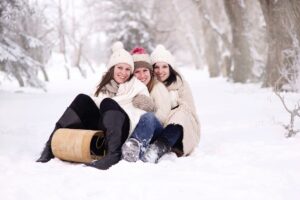 Tres mujeres sonrientes con gorros de lana sentadas en la nieve junto a un trineo de madera