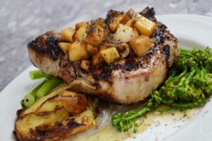 Chuleta de cerdo a la plancha con manzana caramelizada, bimi y patatas asadas en plato blanco