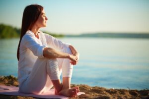 Mujer vestida de blanco meditando sentada en la orilla de un lago al atardecer