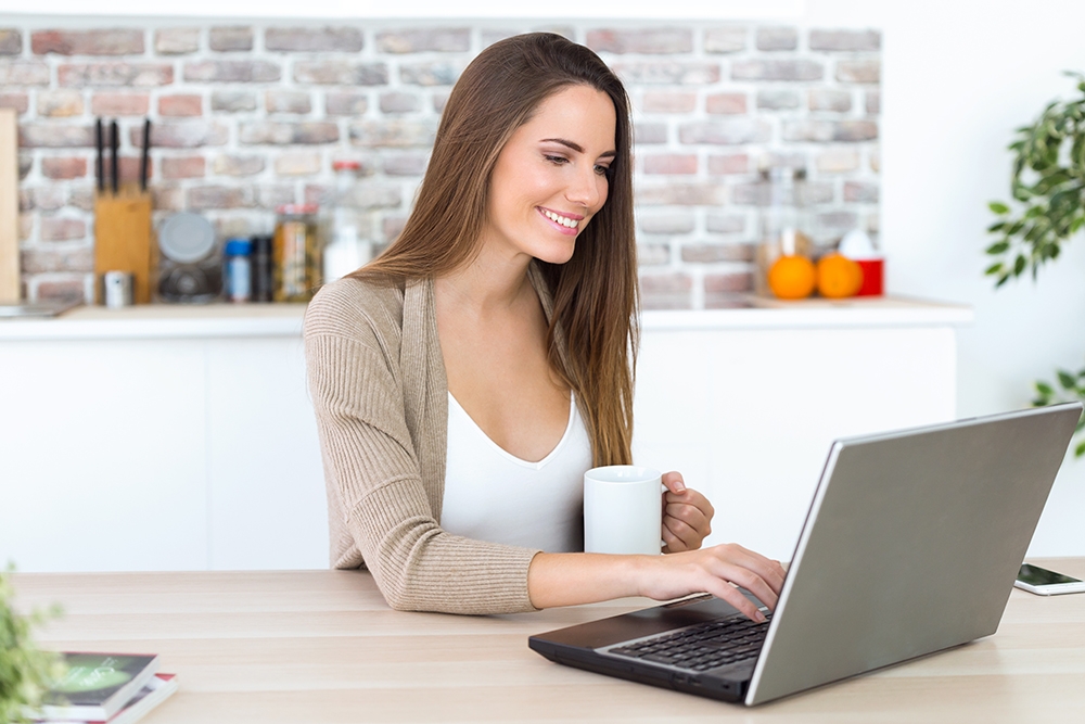 Mujer joven sonriente usando un portátil y tomando café en la cocina de casa