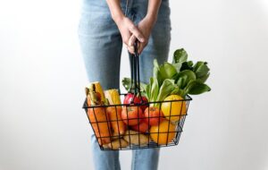Persona en vaqueros sostiene cesta de metal con verduras y frutas frescas sobre fondo blanco