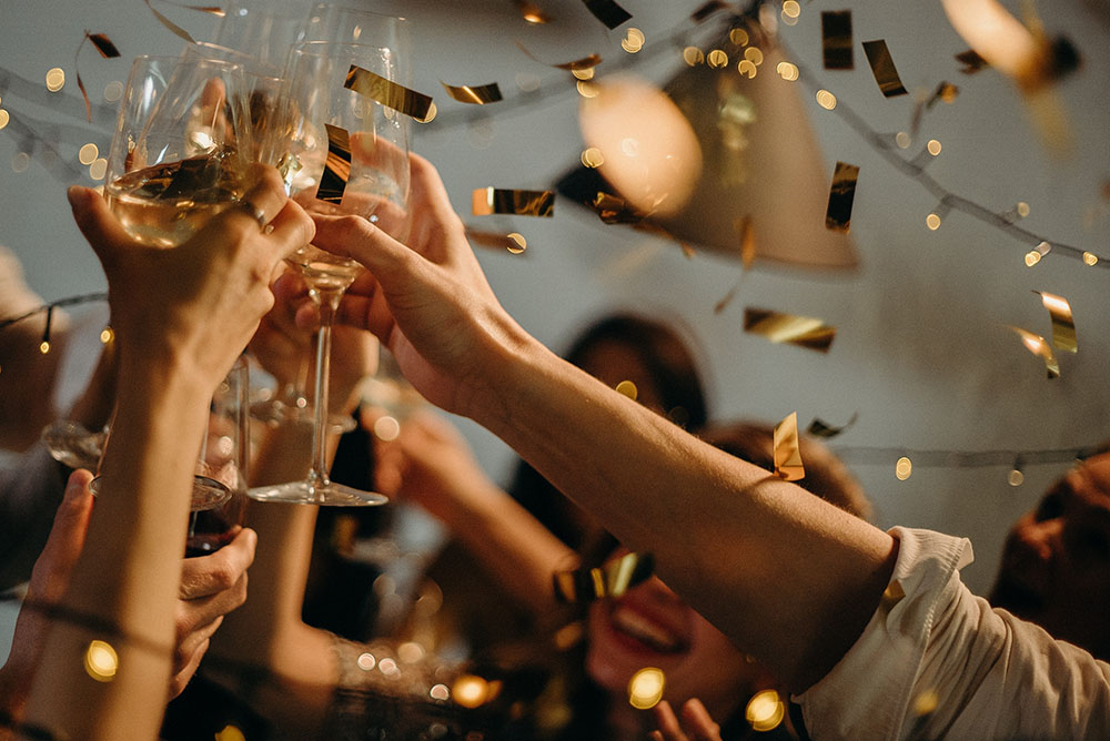 Manos brindando con copas de vino entre confeti dorado y luces cálidas en una fiesta
