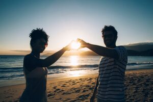 Pareja sonriente formando un corazón con las manos frente al atardecer en la playa
