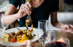 Mujer comiendo verduras asadas con tenedor en un restaurante junto a copas de vino y una vela