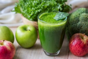 Batido verde detox en vaso rodeado de manzanas, brócoli, espinacas y lechuga sobre mesa de madera