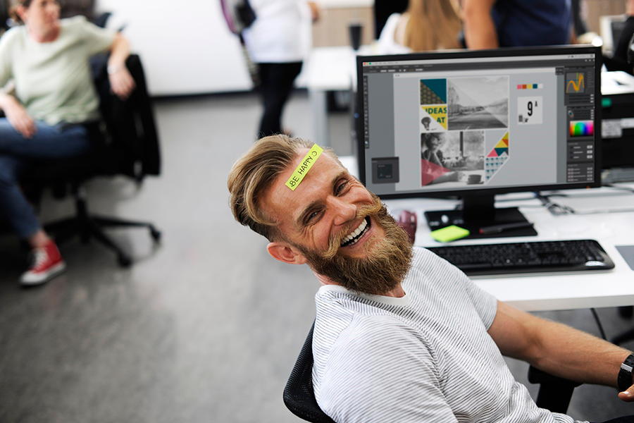 Hombre barbudo sonriente con nota adhesiva Be Happy en la frente en una oficina de diseño