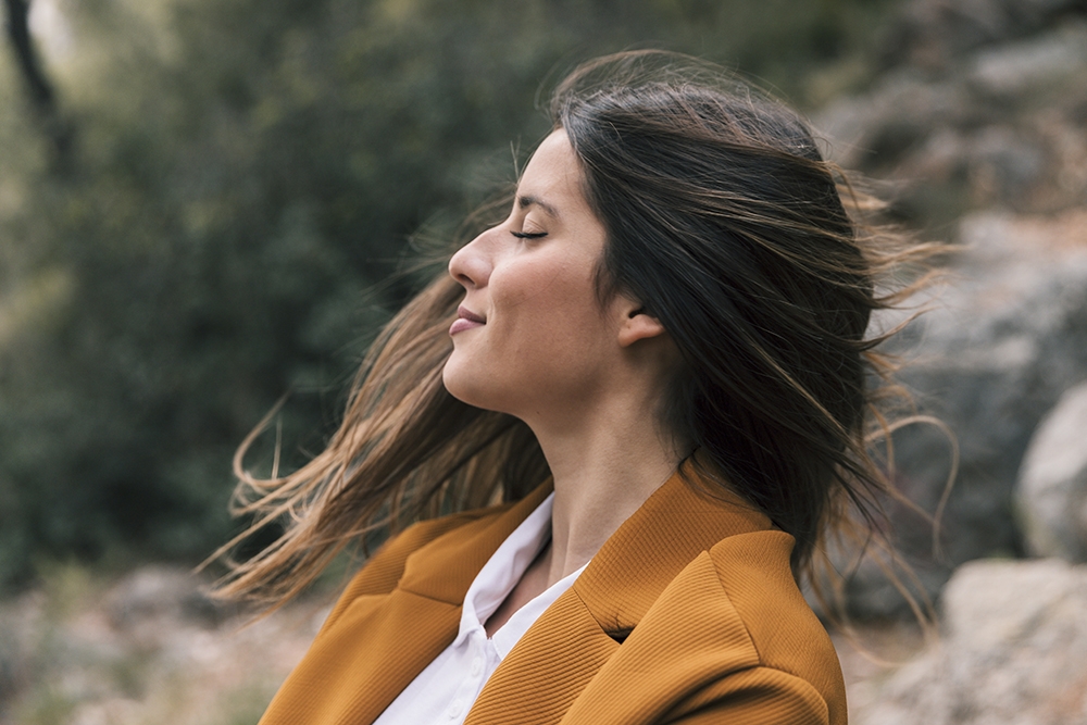 Mujer de perfil con blazer mostaza y ojos cerrados respirando al aire libre con el pelo al viento