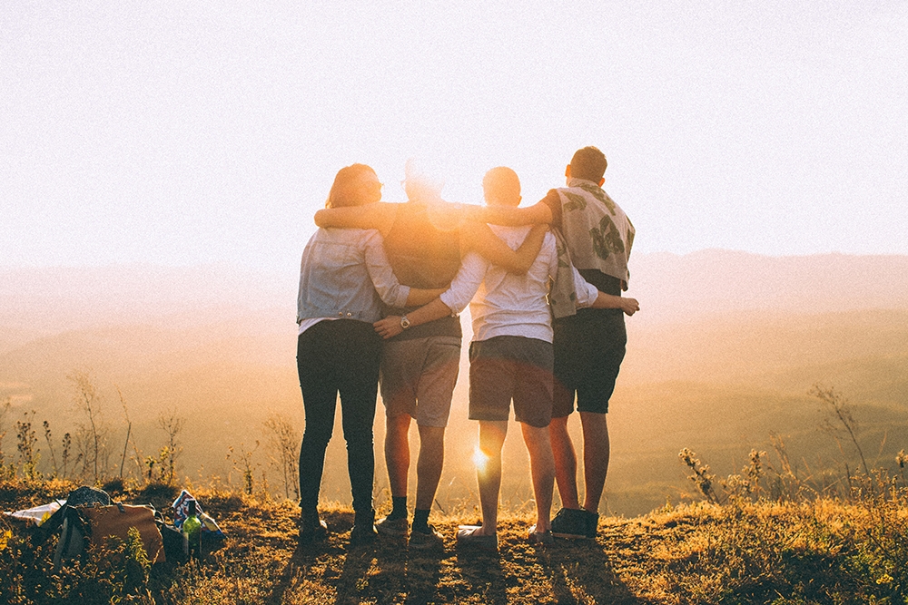 Grupo de cuatro amigos abrazados de espaldas contemplando el atardecer sobre las montañas