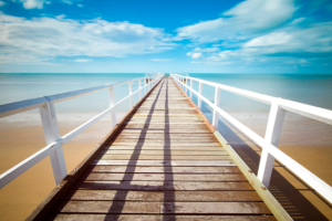 Muelle de madera con barandillas blancas adentrándose en el mar bajo cielo azul con nubes