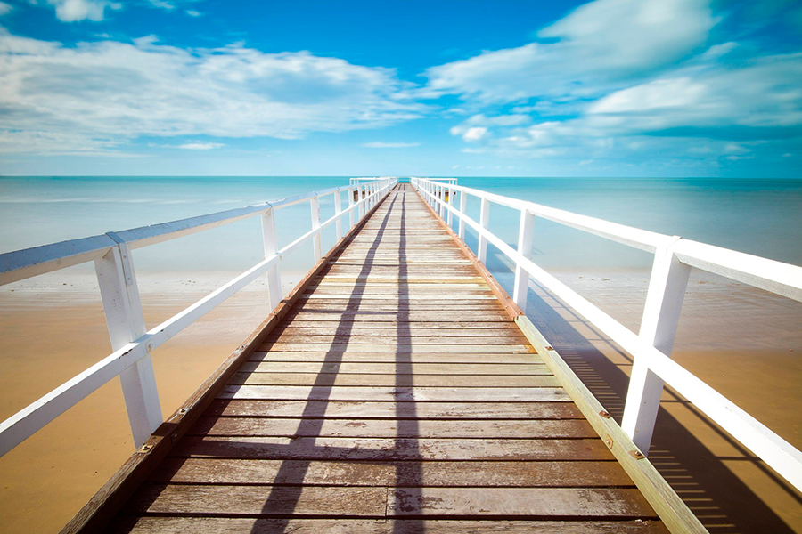 Muelle de madera con barandillas blancas adentrándose en el mar bajo cielo azul con nubes