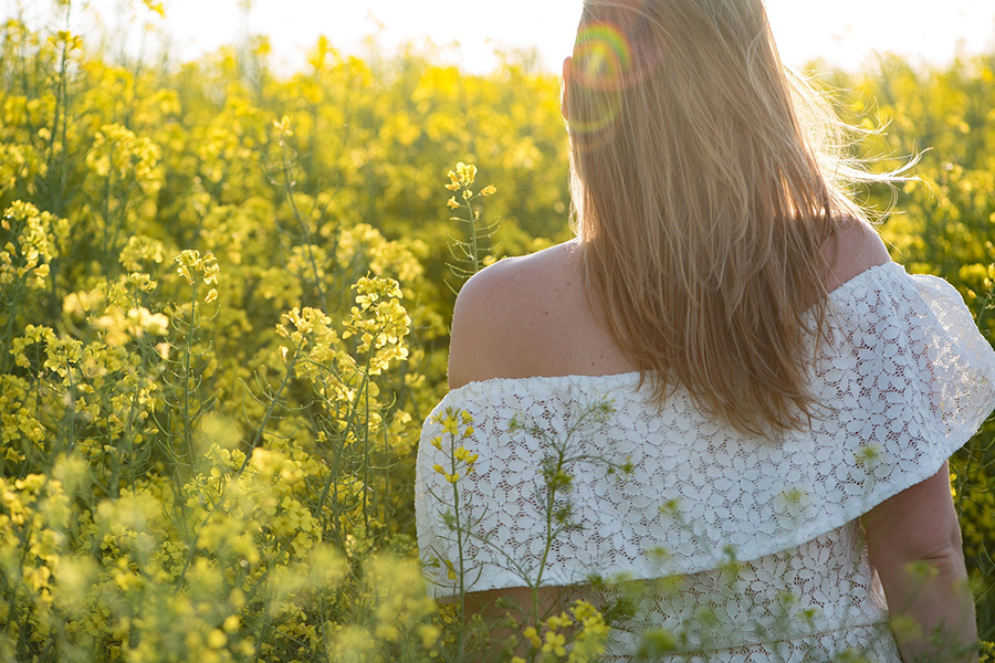 Mujer de espaldas con vestido blanco de encaje en un campo de flores amarillas al atardecer