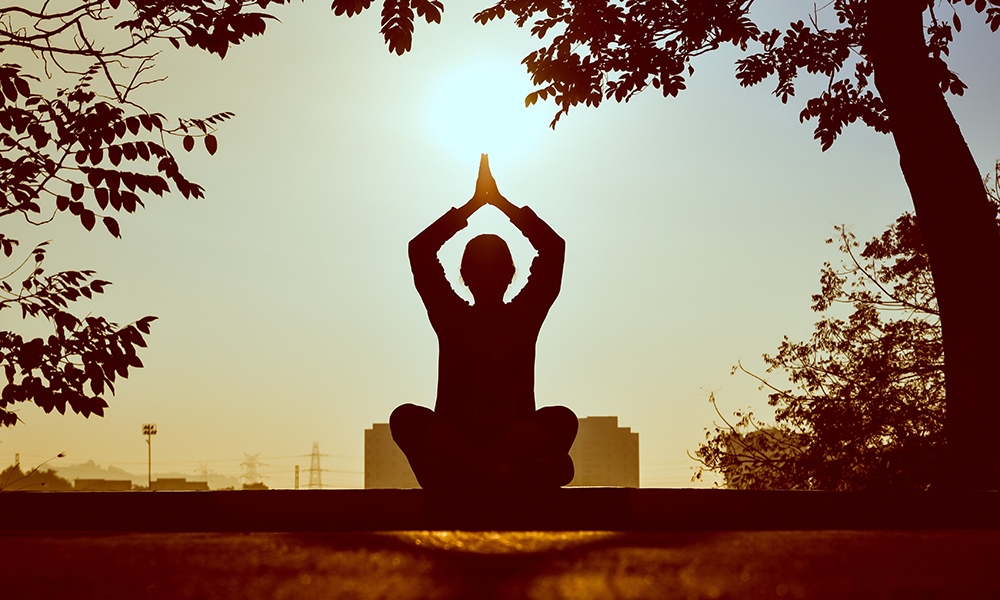 Silueta de persona meditando en postura de yoga con manos juntas al atardecer bajo un árbol