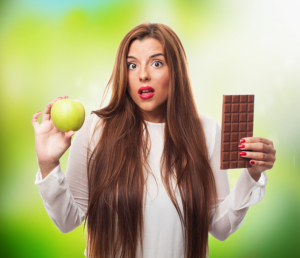 Mujer sorprendida sosteniendo una manzana verde y una tableta de chocolate, dudando entre ambas
