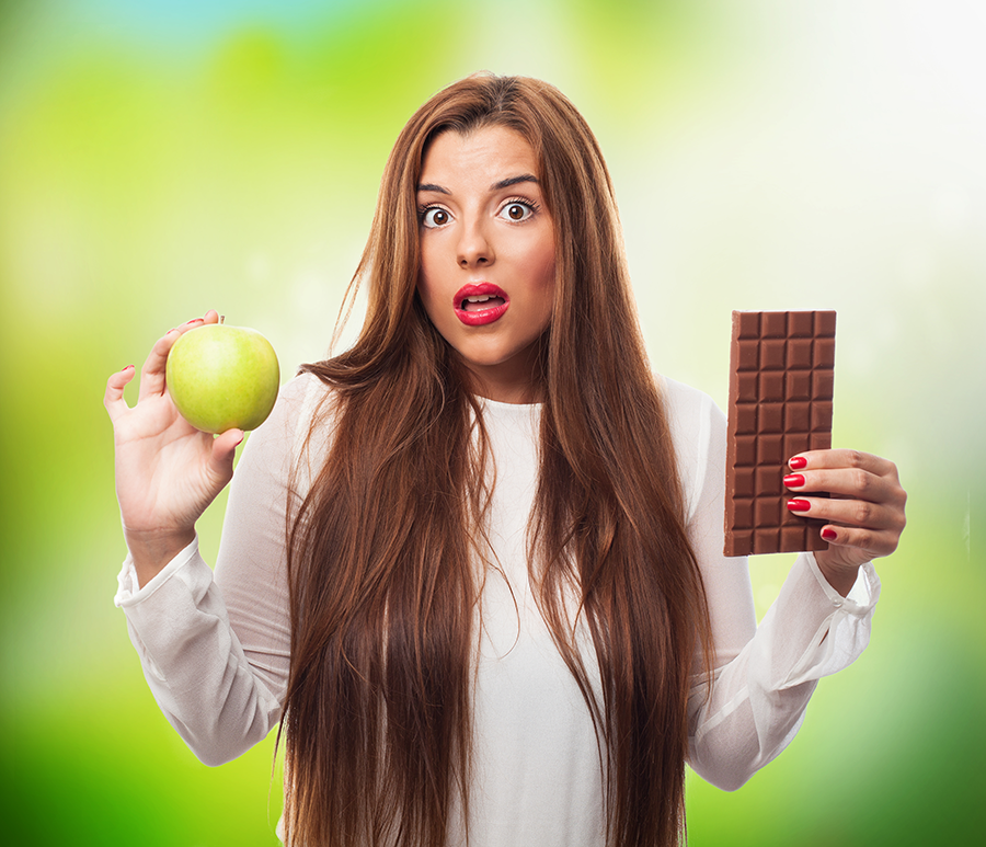 Mujer sorprendida sosteniendo una manzana verde y una tableta de chocolate, dudando entre ambas