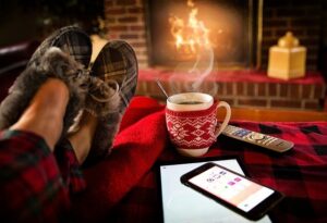 Pies con pantuflas junto a chimenea encendida, taza humeante, manta roja y móvil en un ambiente invernal acogedor