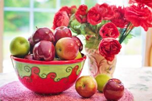 Cuenco verde y rojo con manzanas rojas y verdes junto a ramo de flores rojas en una ventana