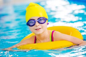 Niña con gorro amarillo y gafas de natación sonríe apoyada en un flotador dentro de la piscina