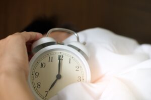 Mano apagando un despertador blanco que marca las siete sobre una cama con sábanas