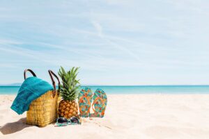 Bolsa de playa con toalla, piña, chanclas y gafas de sol sobre la arena junto al mar
