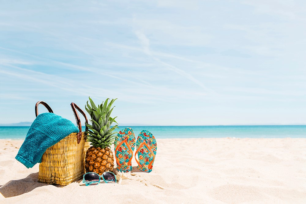 Bolsa de playa con toalla, piña, chanclas y gafas de sol sobre la arena junto al mar