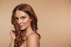Mujer joven pelirroja con melena ondulada, brillante y saludable tocándose el cabello sobre fondo beige