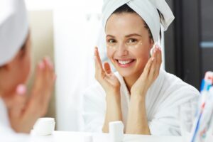 Mujer sonriente con turbante de toalla aplicándose crema facial frente al espejo del baño