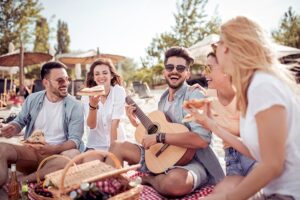 Grupo de amigos riendo en la playa con guitarra, pizza y cesta de picnic en verano