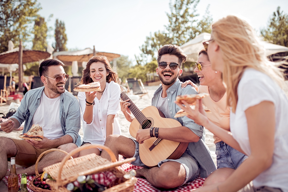 Grupo de amigos riendo en la playa con guitarra, pizza y cesta de picnic en verano