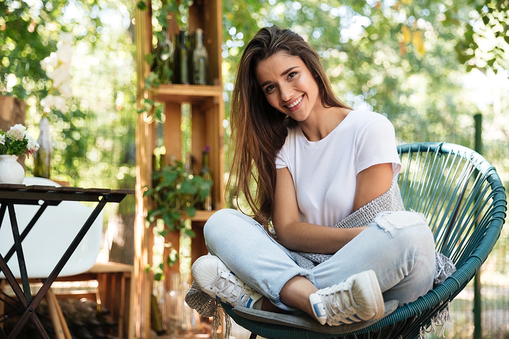 Mujer joven sonriente sentada relajada en una silla de jardín rodeada de plantas en verano