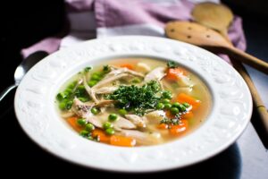 Plato hondo con sopa de pollo desmenuzado, guisantes, zanahoria y perejil junto a cuchara de madera