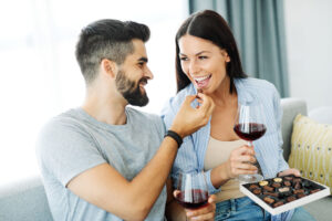 Pareja sonriente disfrutando de bombones de chocolate y copas de vino tinto en el sofá