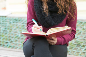 Mujer con jersey granate escribiendo con bolígrafo en una libreta de tapa roja al aire libre