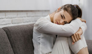 Mujer sentada en el sofá abrazando sus rodillas con expresión de tristeza y preocupación