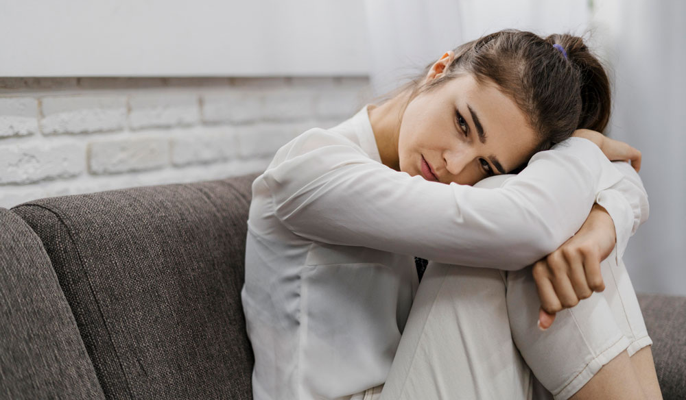 Mujer sentada en el sofá abrazando sus rodillas con expresión de tristeza y preocupación