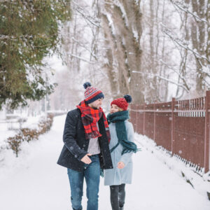 Pareja abrigada con gorros y bufandas caminando por un paseo nevado entre árboles
