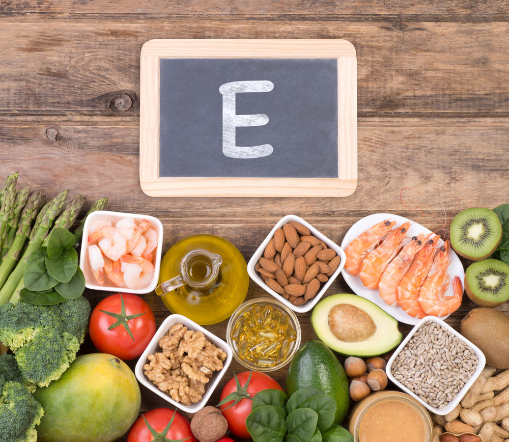 Alimentos ricos en vitamina E como frutos secos, aguacate, aceite, verduras y gambas junto a pizarra con letra E